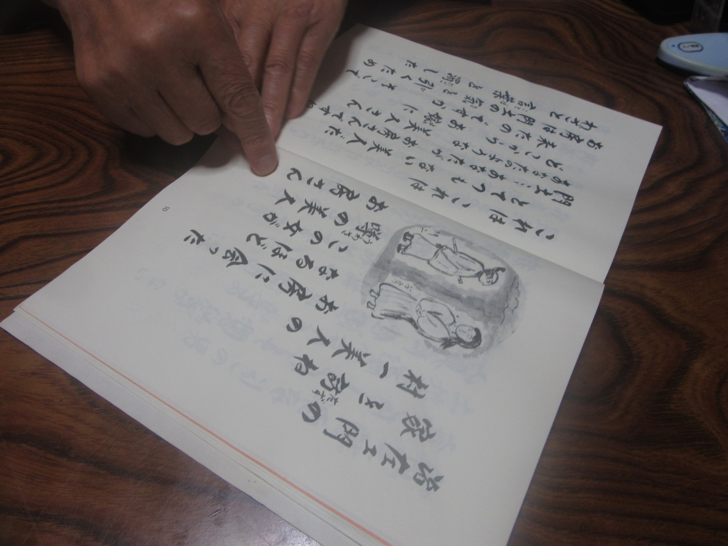 和室の木製テーブルの上で、筆文字風の文章と水墨画風の挿絵が描かれた開いた本のページを、右手の人差し指で指している手元をアップで撮影した写真