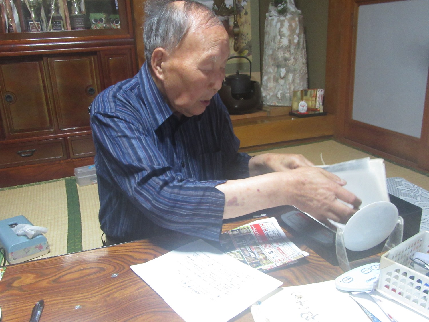 周囲には文房具や日用品が置かれている和室のテーブルの前に年配の小野さんが座り、紙や本、新聞などが入ったケースを整理しながら資料を取り出している様子の写真