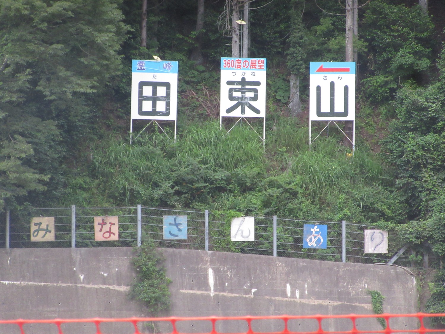 木々に囲まれた山の斜面に「田束山」と一文字ずつ書かれた大きな看板が3つ立っていて、それぞれの看板の上には「霊峰」「360度の展望」「左矢印」の文字があり、下の柵には「みなさんありがとう」と一文字ずつ書かれたひらがなのパネルが並んでいる写真