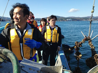 船の上でライフジャケットを着た数人の視察者たちが笑顔で牡蠣養殖の様子を見学している写真