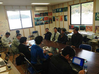 会議室のような部屋で複数人がテーブルを囲んで話し合いやパソコン作業を行っている写真