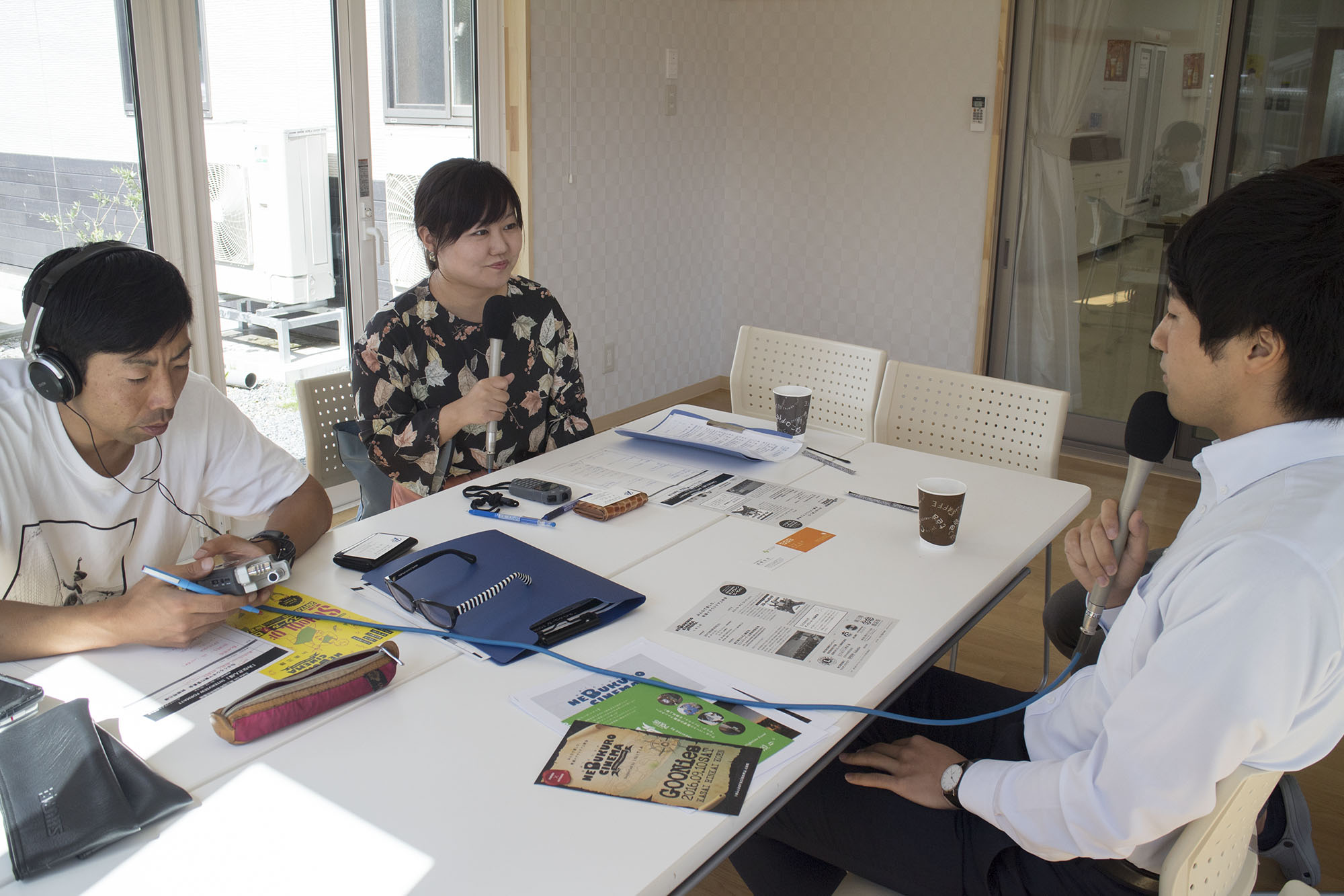 白いテーブルを囲んで、ヘッドホンをつけて機材を操作する男性、マイクを持って話を聞く女性リポーター、インタビューに答える白いシャツの阿部将己さんの三人が座っている収録中の写真