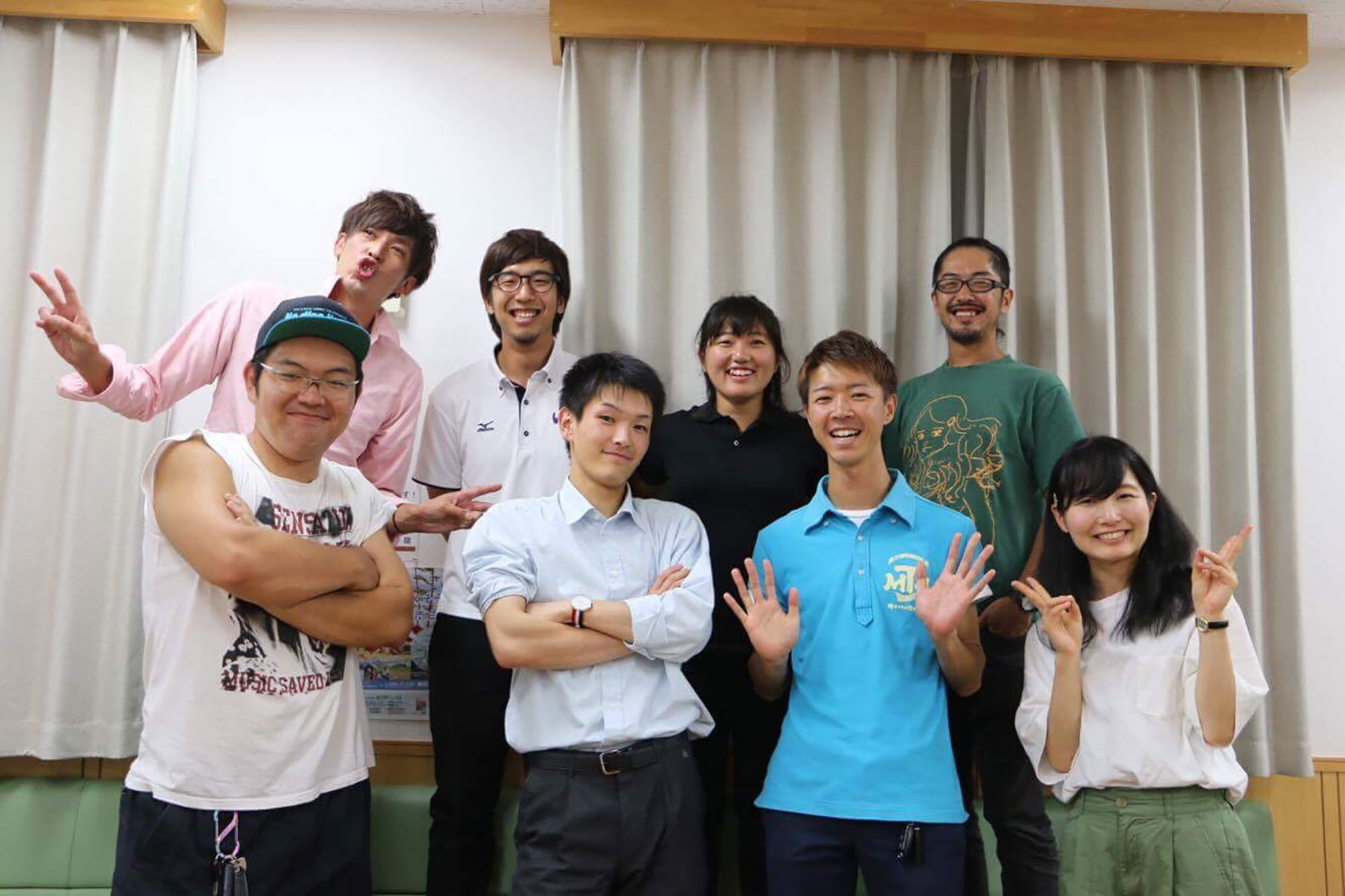 室内で、男女あわせて8人の若者が肩を寄せ合いながら明るい笑顔でピースサインなどのポーズをとっている集合写真