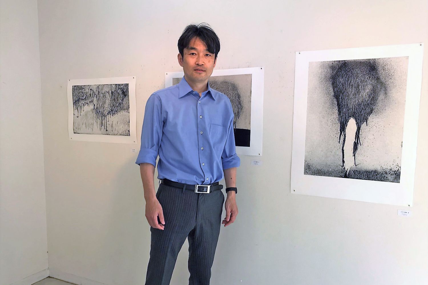 白い壁に展示された3点の抽象的な白黒の作品の前に、青いシャツと灰色のズボンを着た谷村さんが立っている写真