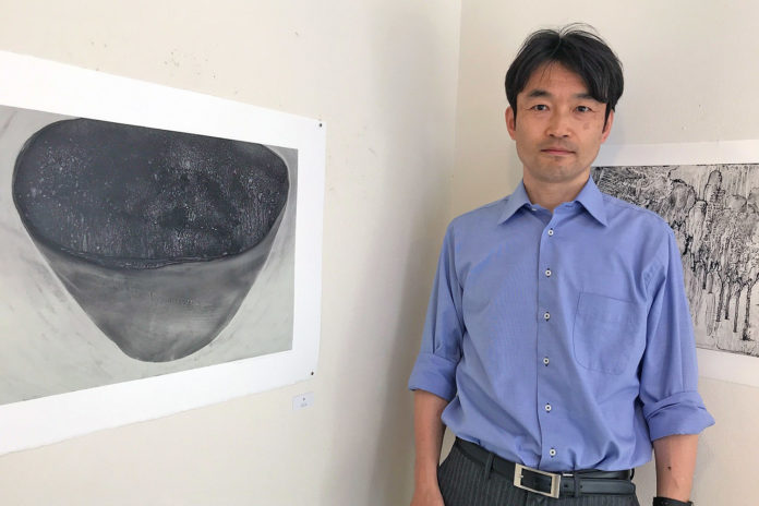 白い壁に黒い器の絵が飾られている横に、青いシャツを着た谷村さんが立っている写真