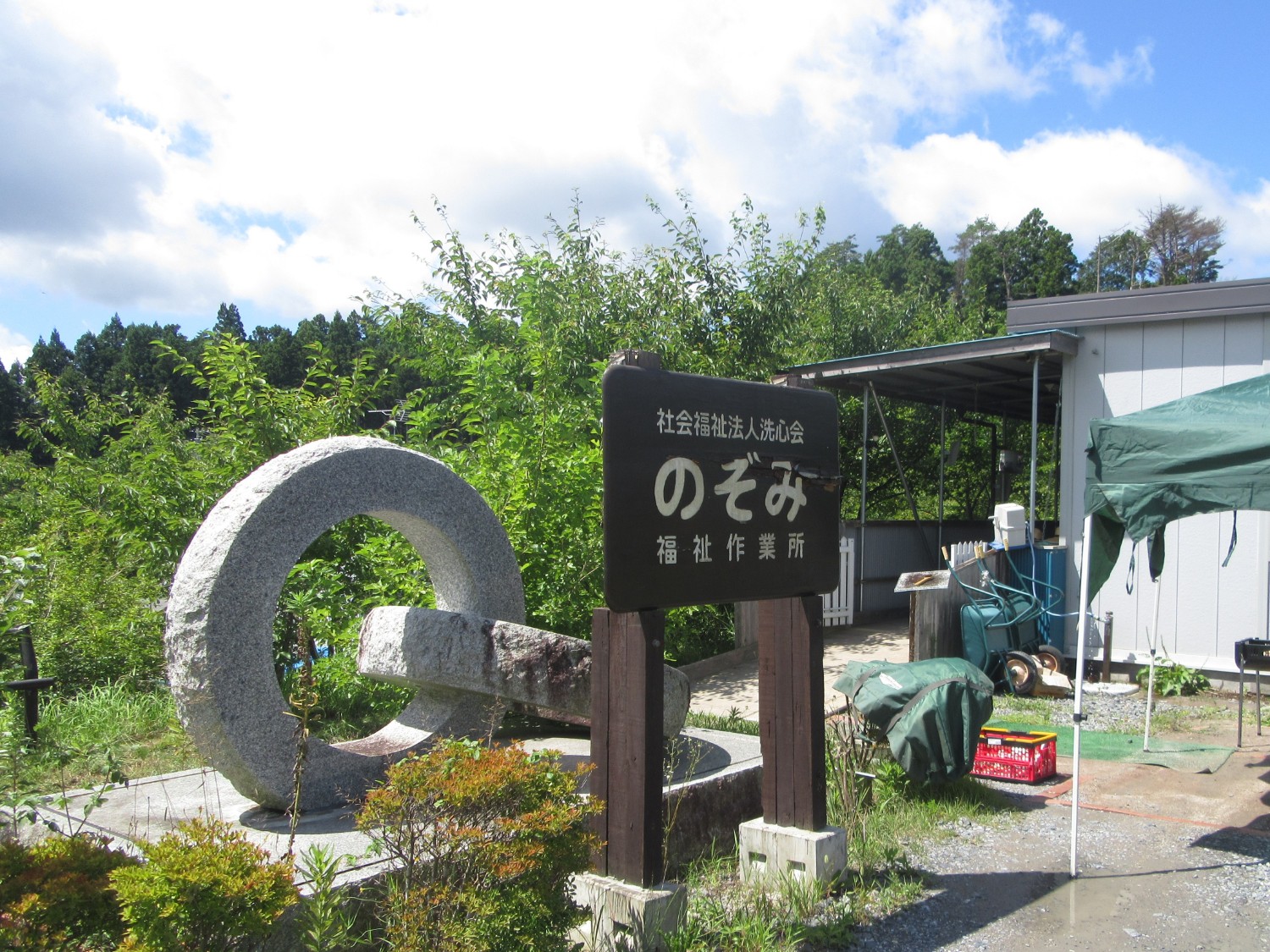 緑に囲まれた場所に「のぞみ福祉作業所」の看板と石のモニュメントが立っている写真