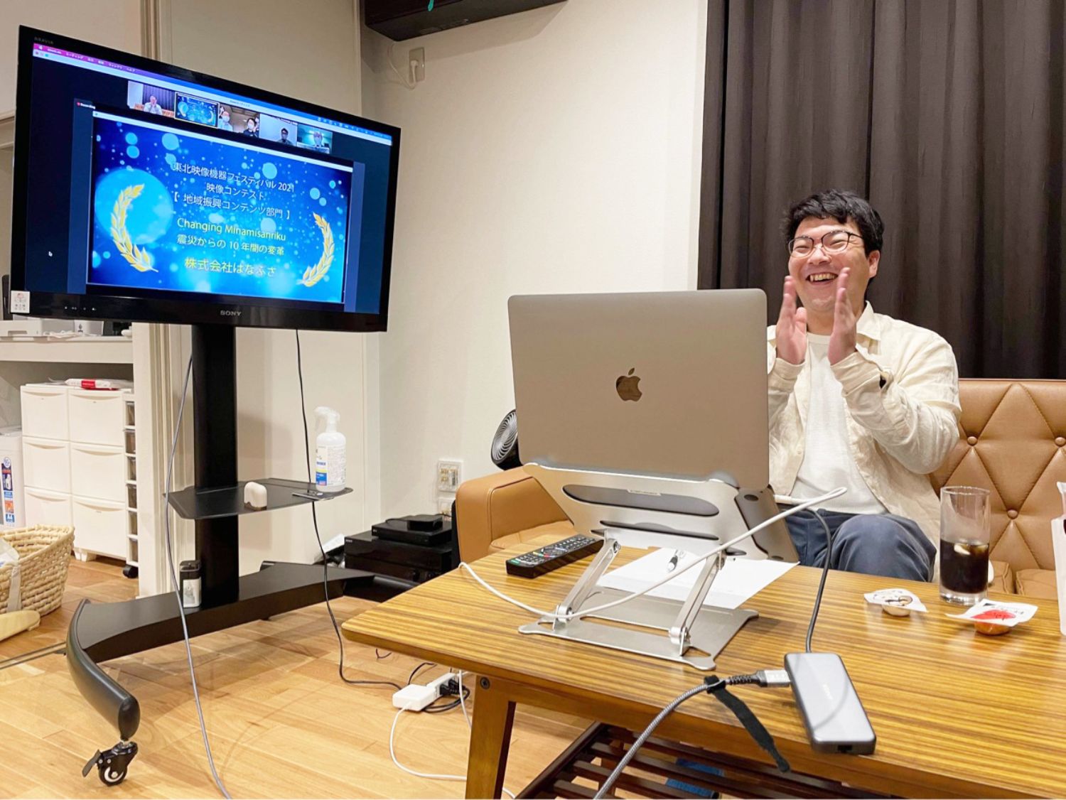 リビングのような室内で、白いシャツを着た佐藤さんがノートパソコンの前に座り、笑顔で拍手をしており、左側の大型モニターには「映像コンテスト受賞」の画面が表示されている様子を写した写真