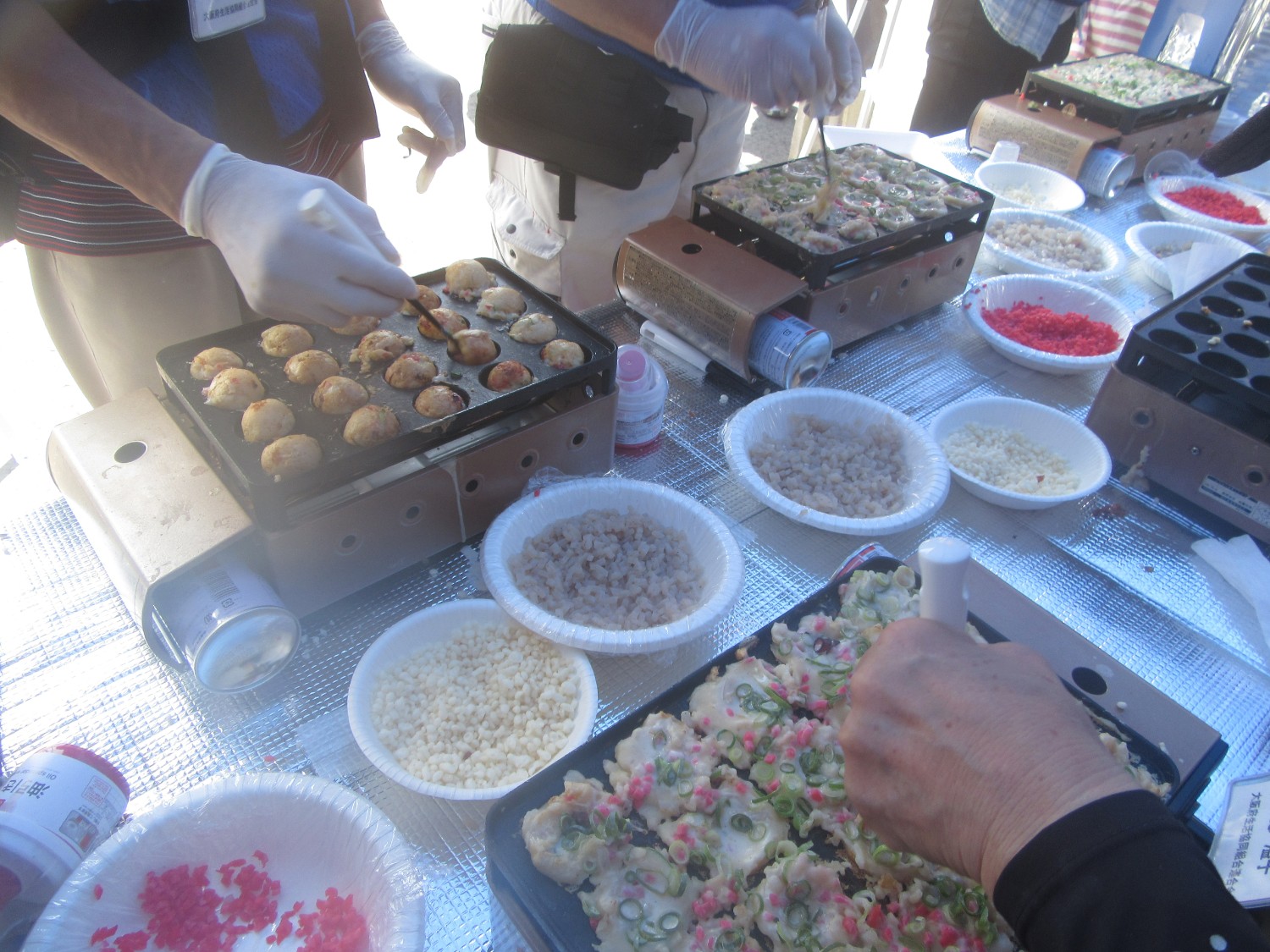 ガス式のたこ焼き機が5台と紙皿に材料が準備され、生地を金串でクルクルと手際よく丸めていく様子の写真