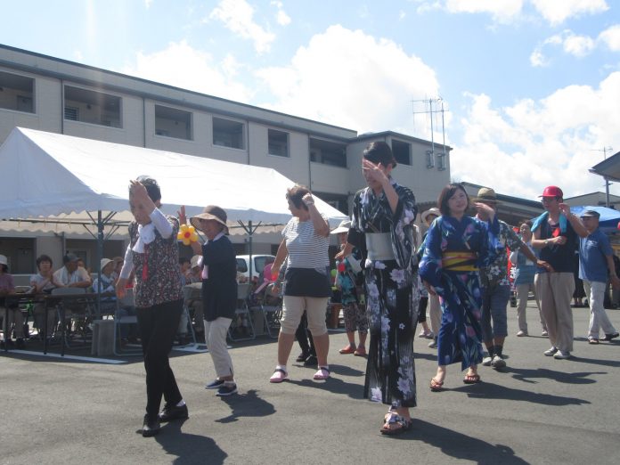 青空の下、左側に白いテントが設置され、浴衣を着た女性や住民の方々が踊りを踊っている様子の写真