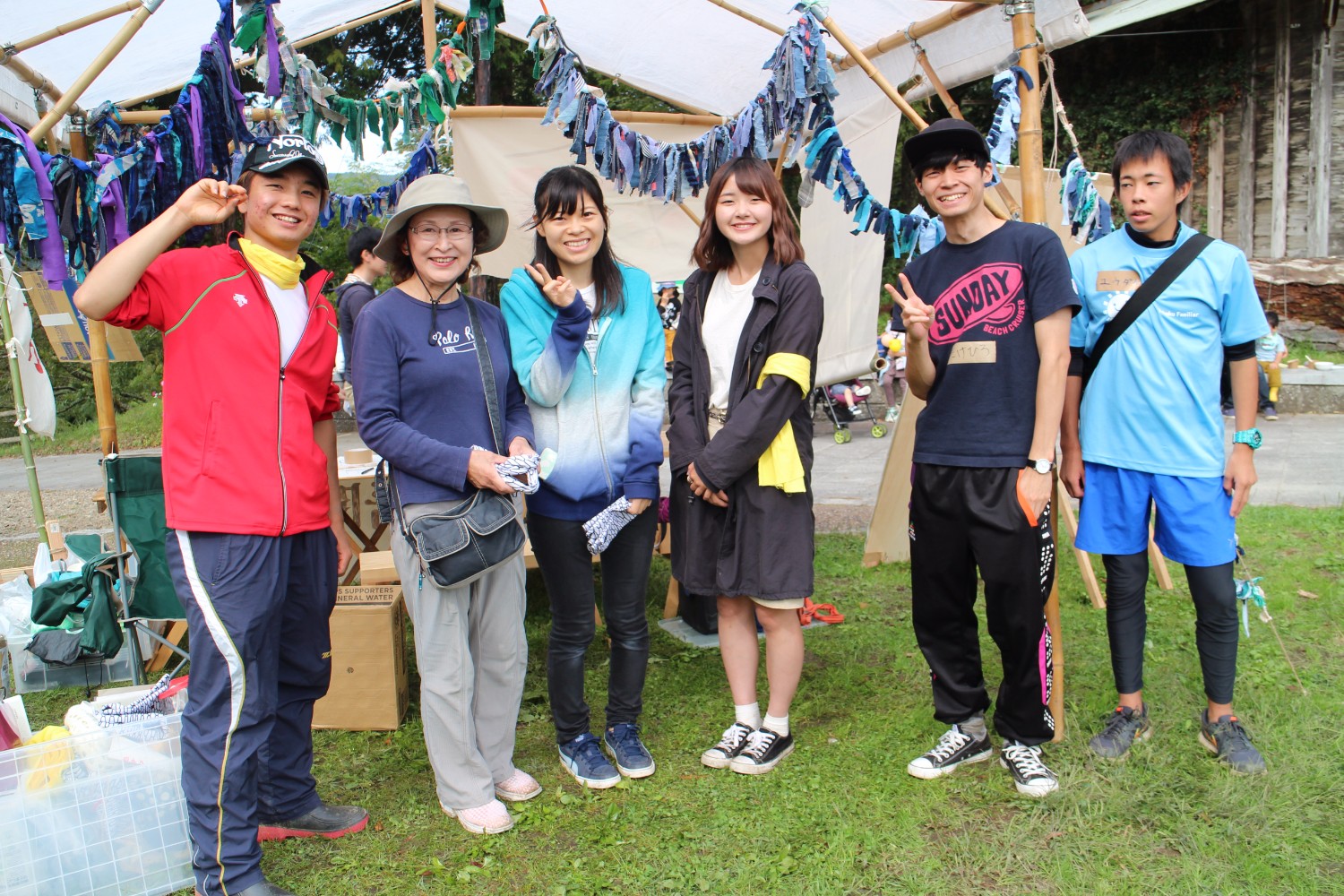 芝生の広場に設置されたテントの木の骨組みに青や紫の布で作られた飾りが取り付けられ、その前で男女の学生や関係者の女性6人が笑顔で並んでいる記念写真