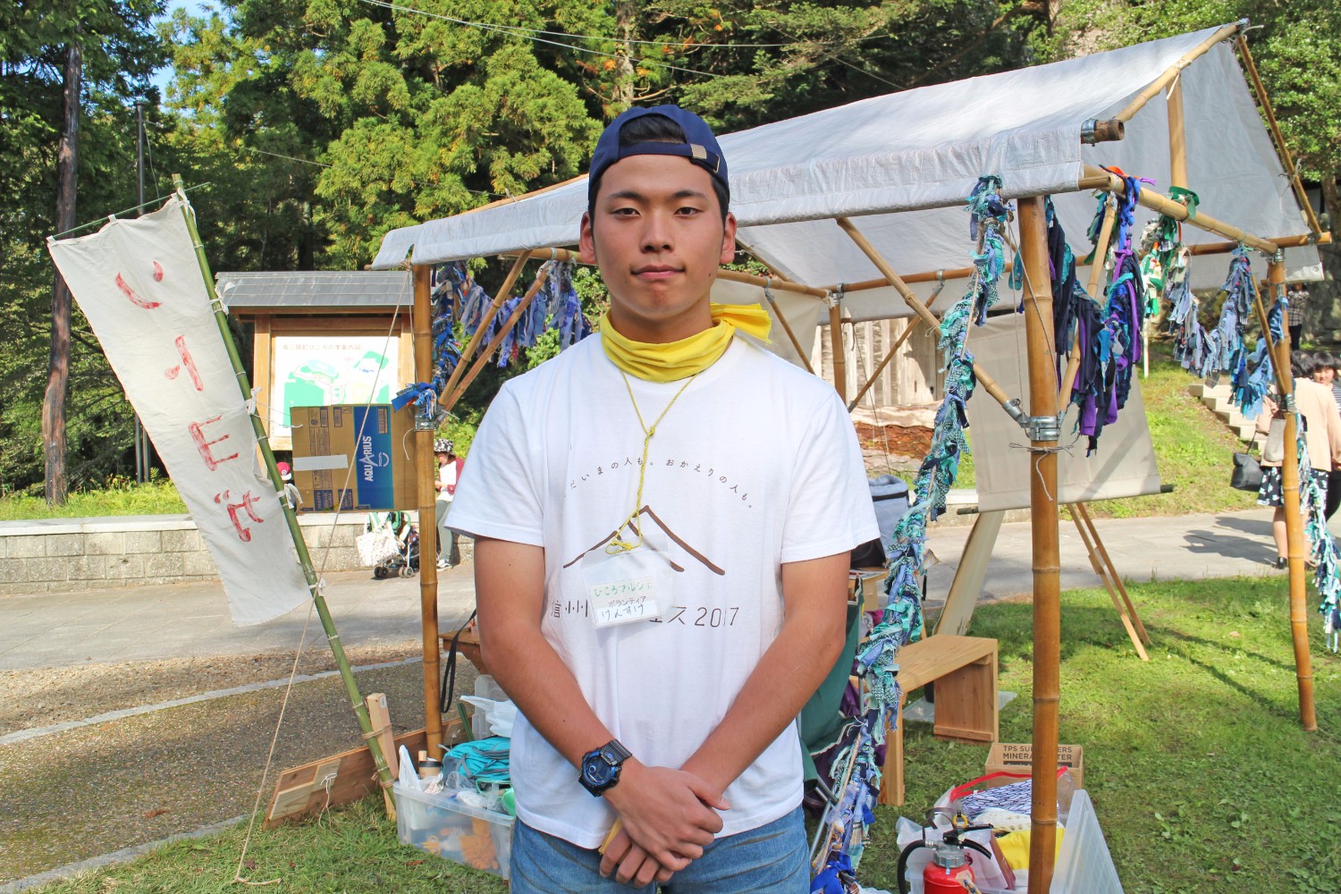 木の骨組みに青や紫の布の飾りが取り付けられた白いテントの前で、姿勢を正して正面を向いて立っている小坂さんの写真