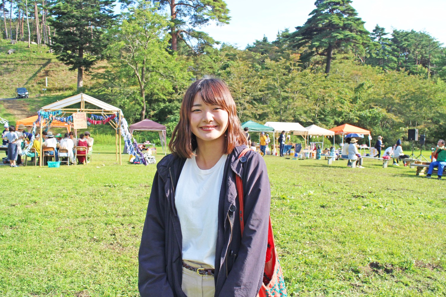 たくさんのテントが立ち並び、ベンチで休憩する人々が見られる芝生の広場に立ち、カメラに笑顔を見せている若井さんの写真