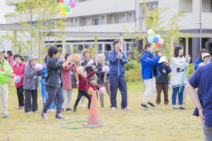 高齢者から若者まで幅広い年齢層の参加者がグラウンドに並び、カラフルなポンポンを持って拍手や応援をしている様子で、地域の運動会の応援風景の写真