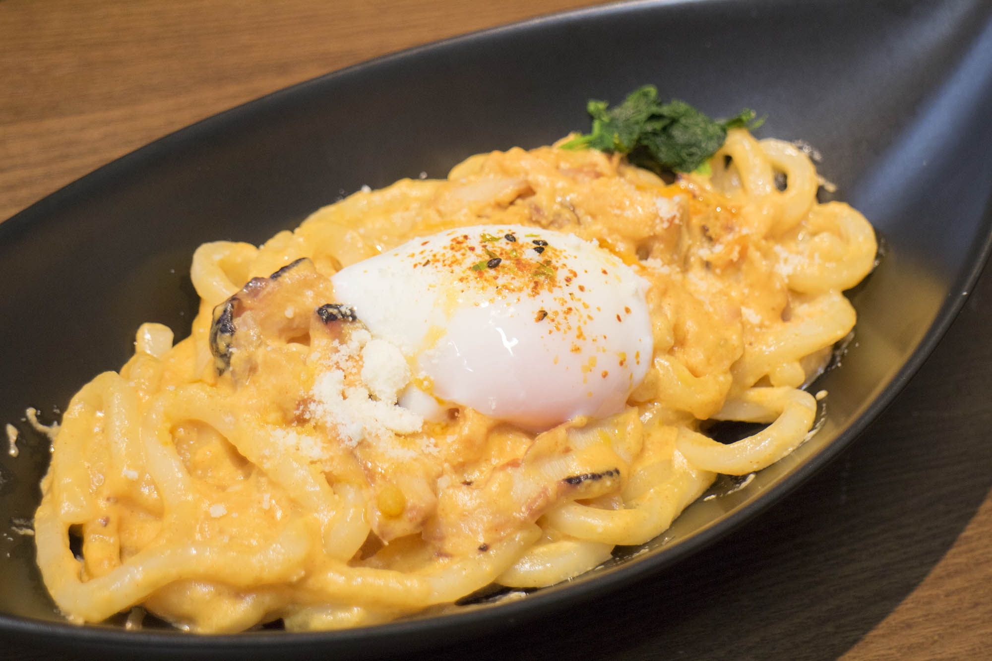 黒い楕円形の皿に盛られた濃厚なクリームソースうどんの上に温泉卵がのっている料理のアップ写真