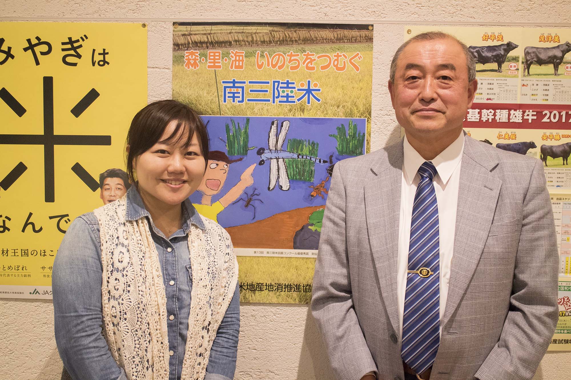 「南三陸米」などのポスターが貼られた壁の前で、リポーターの女性とグレーのスーツを着た阿部さんが並んで立ち、笑顔でカメラを見ている写真