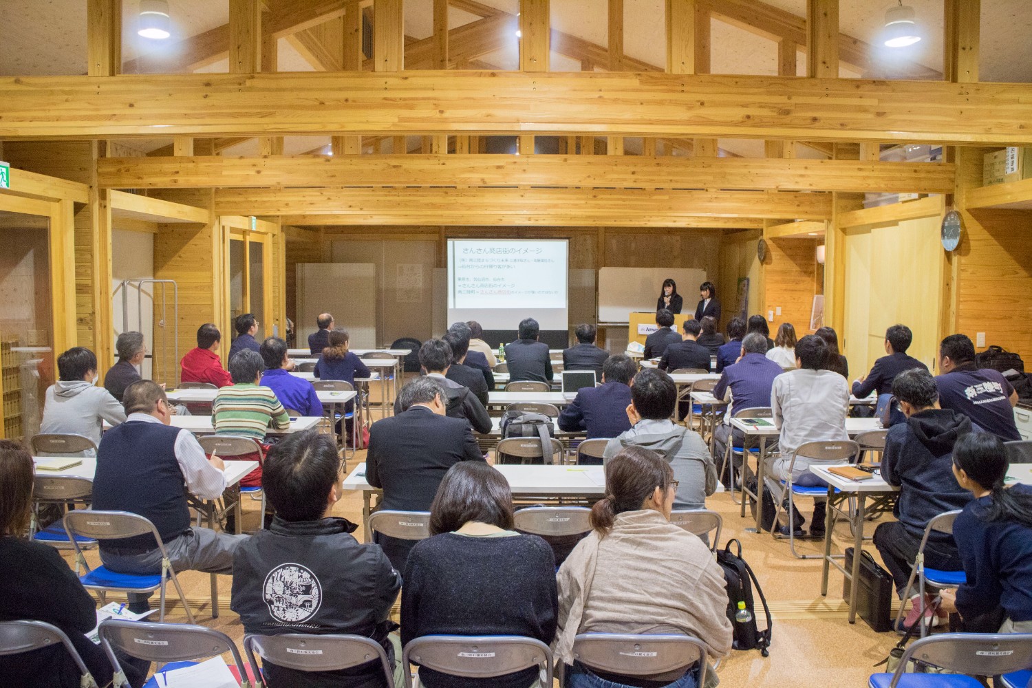 天井が高く、木の梁が見える広い会場で、たくさんの出席者たちが席に座り前方のスクリーンを見ている様子を後方から撮影した写真