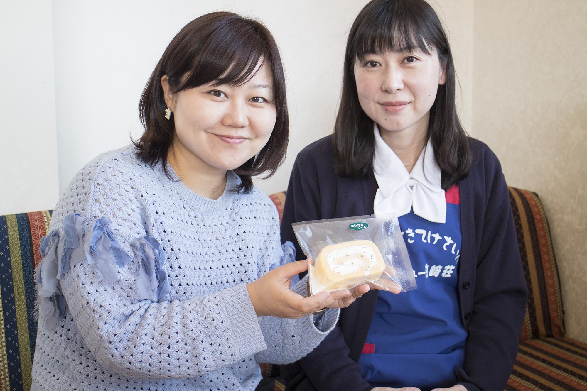 女性リポーターが透明な袋に入ったロールケーキを両手で持ち、隣に座った佐藤さんと笑顔で並んで写っている写真