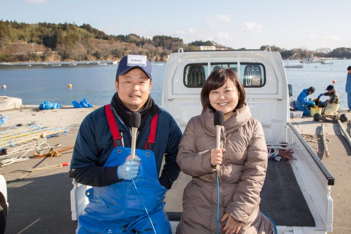 漁港を背景に白い軽トラックの荷台に並んで腰掛け、笑顔でマイクを持っている高橋さんと女性リポーターの写真