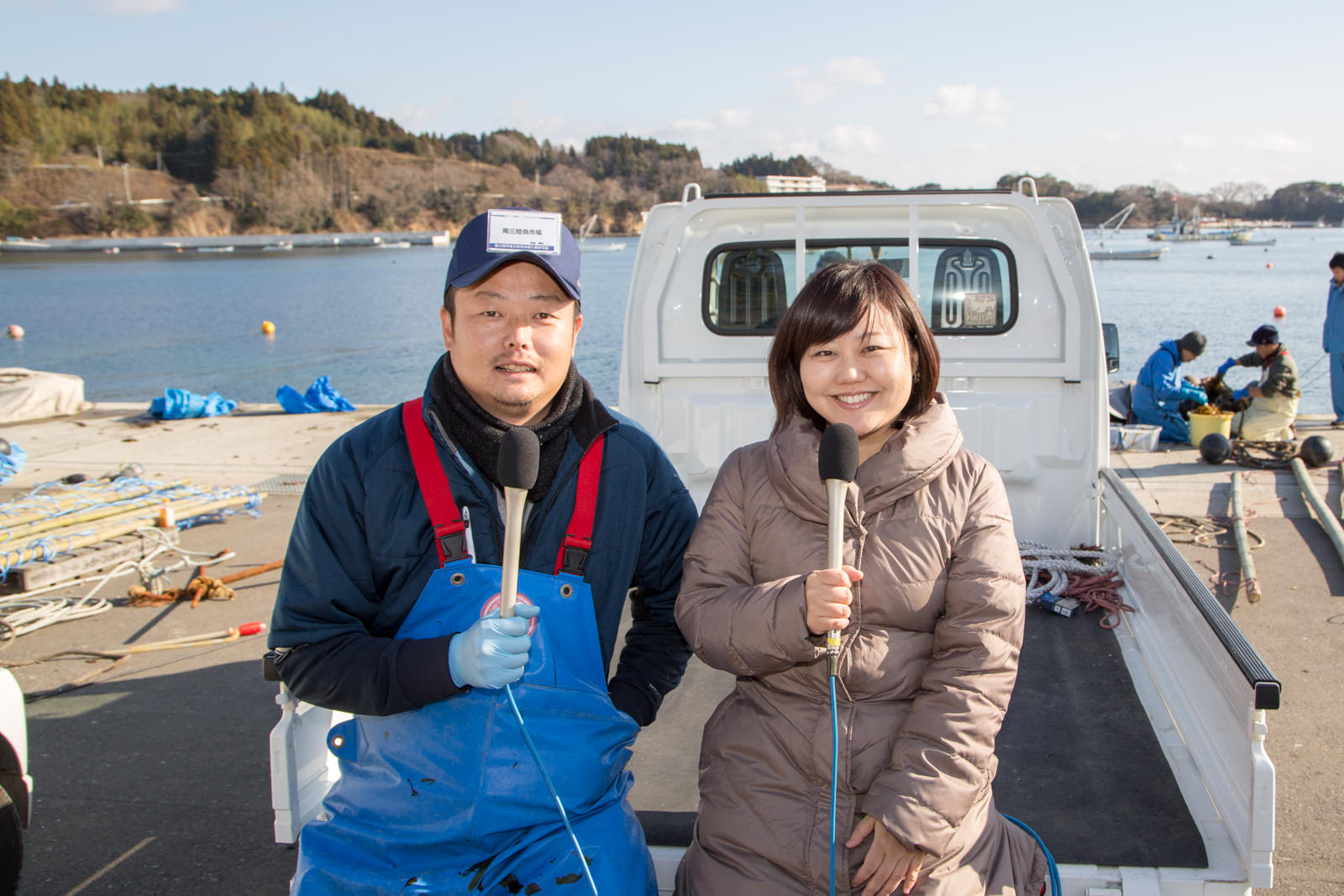 漁港を背景に白い軽トラックの荷台に並んで腰掛け、笑顔でマイクを持っている高橋さんと女性リポーターの写真