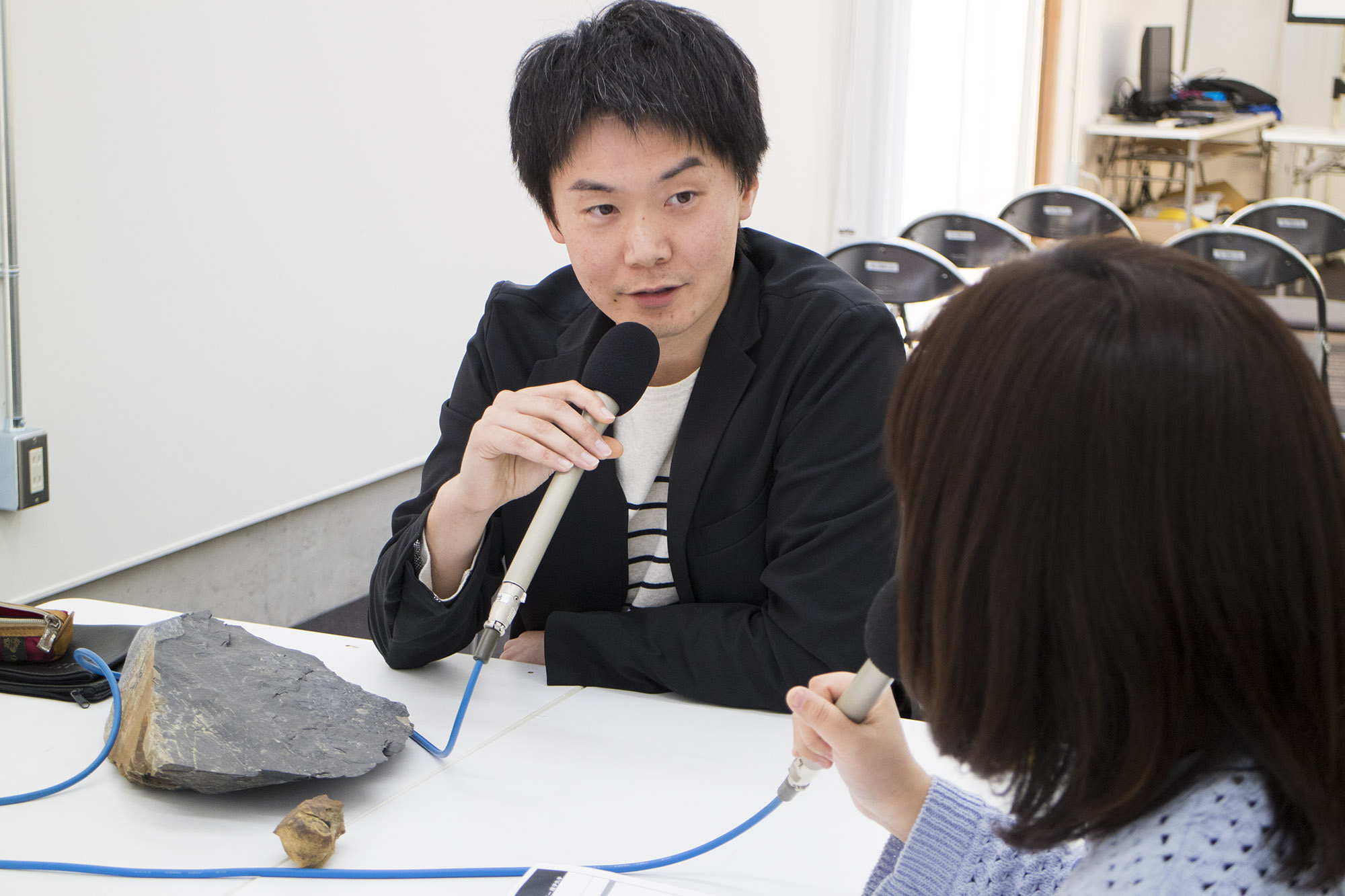 マイクを持った大森さんと女性リポーターが、大きな石とアンモナイトの化石がおかれたテーブルをはさんで対談している写真