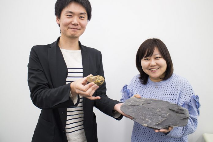 黒いジャケット姿の大森さんがアンモナイトの化石を手に、女性リポーターと一緒に大きな石を持って笑顔を見せている写真