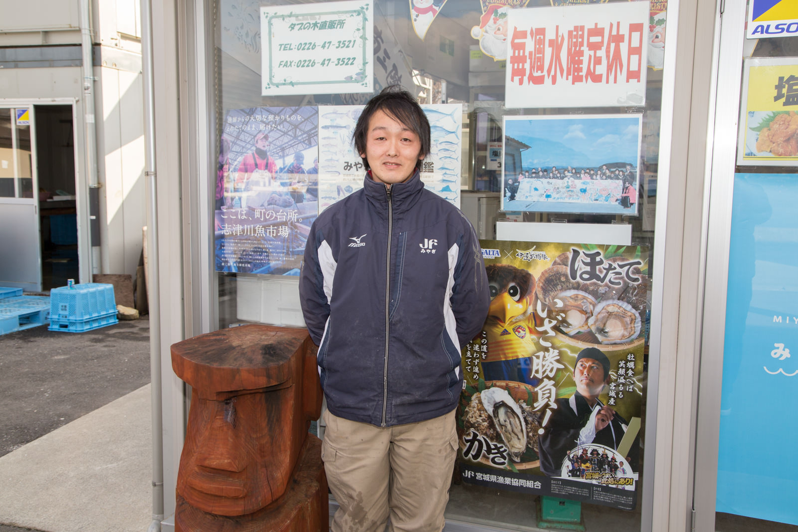 たくさんのポスターなどが貼られたガラス窓の外で、大きな木彫りのモアイ像の横に立つ宇野さんの写真