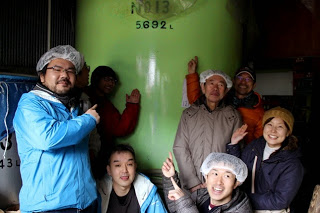 酒蔵の大きなタンクの前で関係者の人たちが笑顔で記念撮影をしている写真