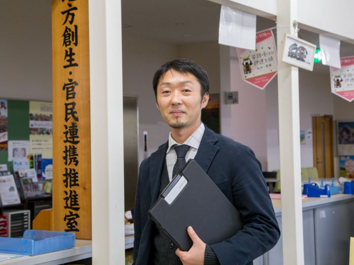 「地方創生・官民連携推進室」と書かれた木製の看板と、事務用の机や掲示物などが並んでいる職場の前で、書類ケースを胸に抱えて立っている男性の写真
