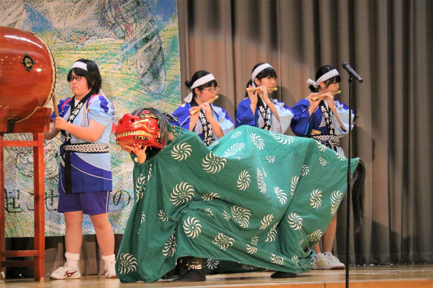 舞台上で太鼓をたたく少女と、横に並んだ笛を吹く少女たちの前で、赤い顔の獅子頭と緑の布をかぶった獅子舞が身をかがめている写真