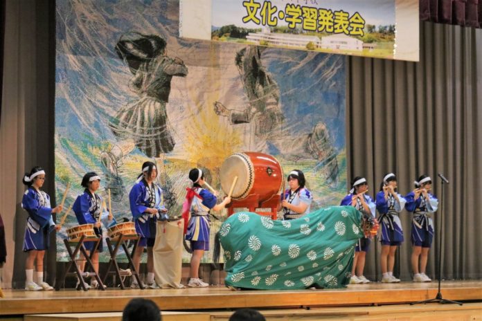 「文化・学習発表会」と書かれた体育館の舞台の上で、法被姿の生徒たちが演奏する太鼓や笛の音色に合わせて、緑の布をかぶった獅子舞が中央で踊っている文化発表会の写真