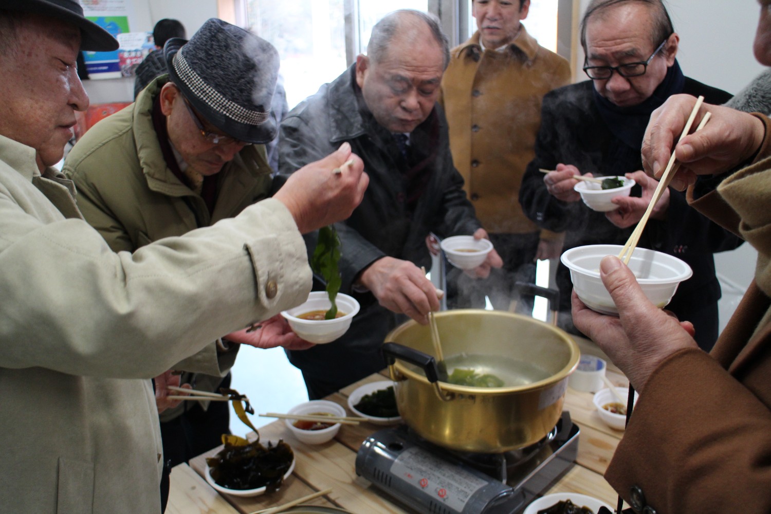 室内のテーブルの上に置かれた鍋でわかめを茹で、周囲の高齢男性たちが小さな器に箸でわかめを取って食べている様子の写真