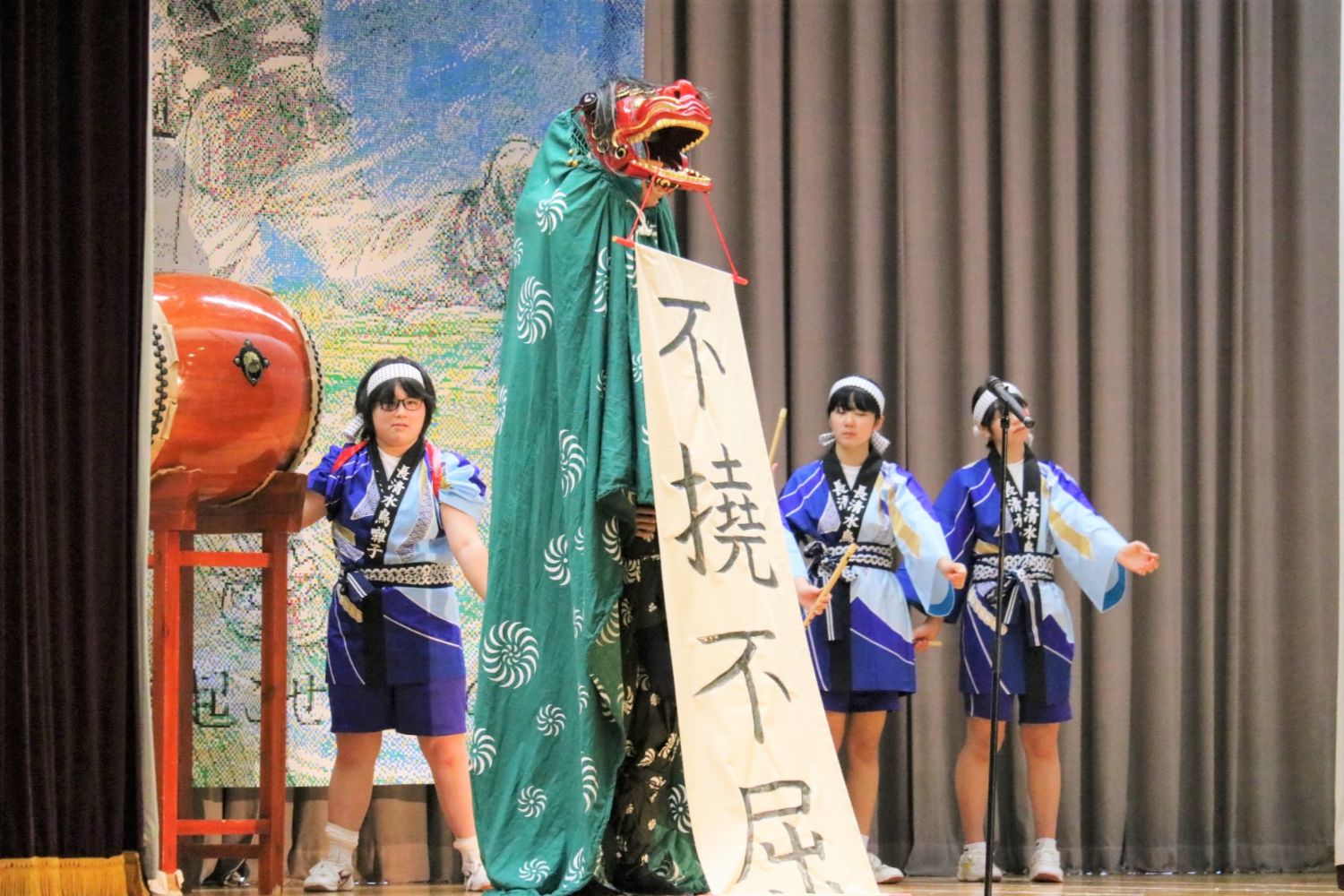 舞台の中央で緑の布をかぶった獅子舞が「不撓不屈」と縦書きで書かれた白い垂れ幕を口にくわえて掲げ、周囲に法被姿の子どもたちが両手を前に出して並んでいる写真