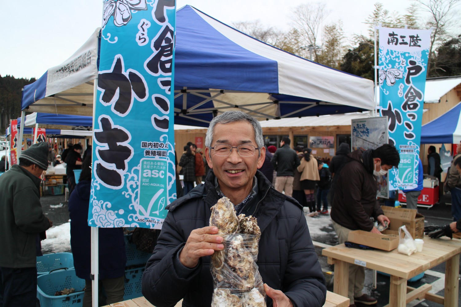 牡蠣販売の屋台の前で、男性がビニール袋に入りきらないほどの大量の殻付き牡蠣を両手で持って笑顔を見せている様子の写真
