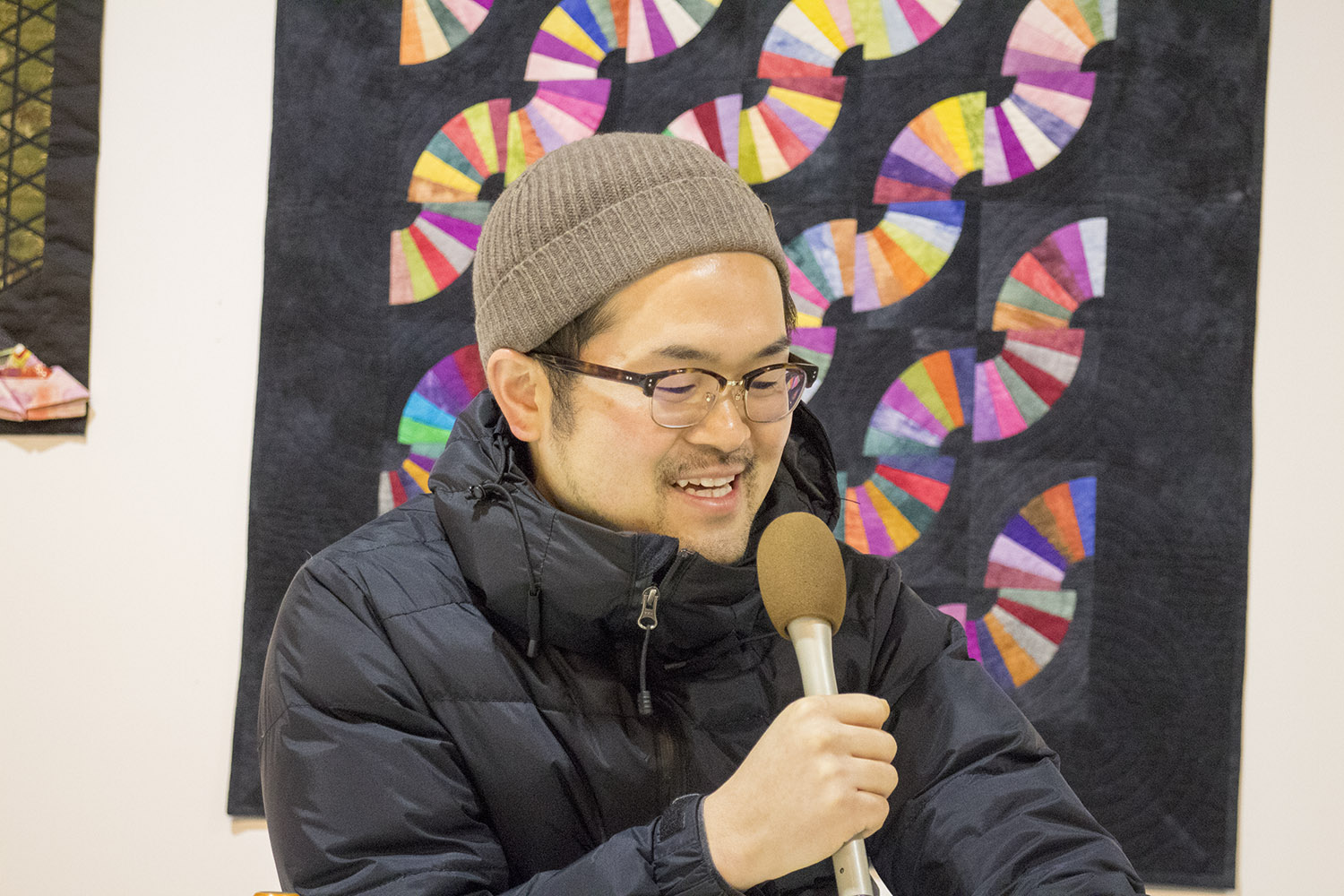 黒い生地に扇形のカラフルなパッチワークが施された作品が飾られた壁を背に、マイクを持って笑顔で話をしている柳井さんの写真