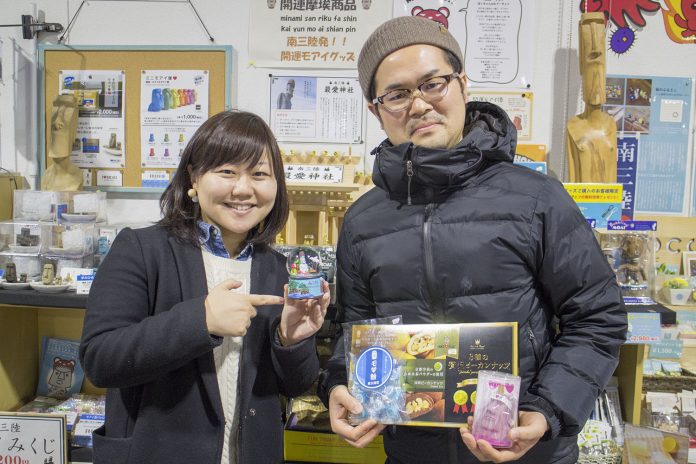 いろいろなモアイグッズが陳列された棚の前で、スノードームや箱入りのお菓子などを手に持った女性リポーターと柳井さんが並んで立っている写真