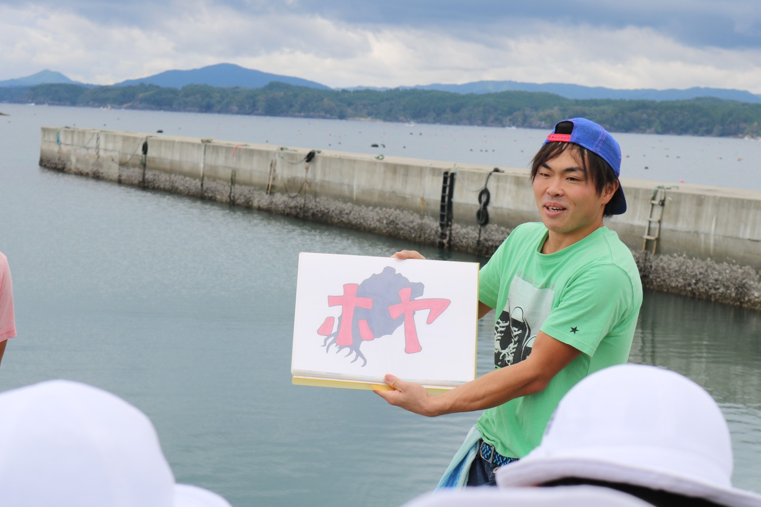 海辺で帽子をかぶった子どもたちに向かって、緑色のシャツを着た男性が「ホヤ」という文字とイラストが描かれたボードを見せながら話している様子の写真