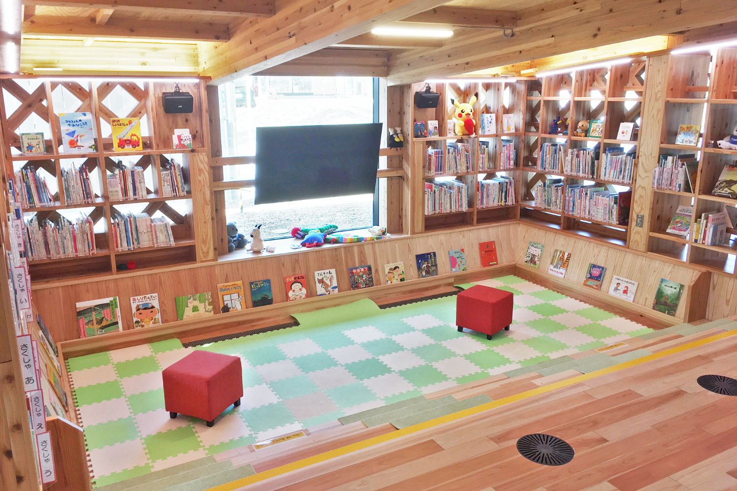 四方を木製の本棚に囲まれ、太陽の光が差し込む部屋の中央に緑と白のマットが敷かれ、2つの赤いスツールが置かれている子ども向け読書スペースの写真