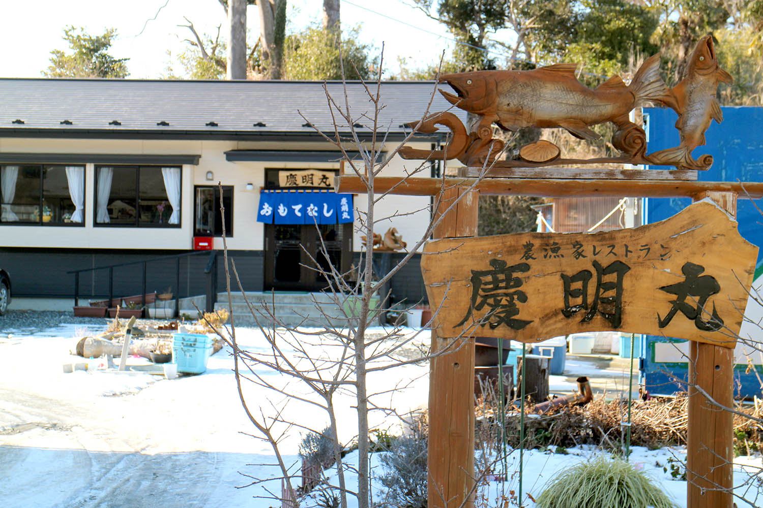入口に青い暖簾がかかった店の建物の前のスペースには雪が積もり、手前に魚の彫刻が施された「慶明丸」と書かれた木の看板が立っている写真