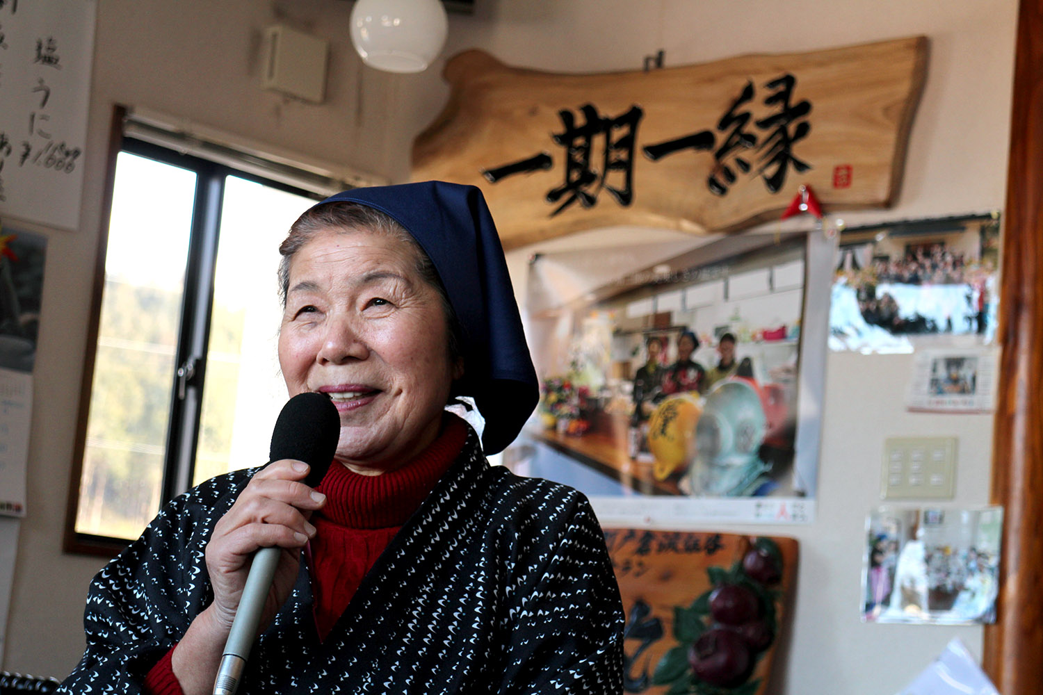 「一期一縁」と書かれた木の看板や大きな写真などが飾られた壁を背景に、マイクを持って笑顔を見せている三浦さんの写真