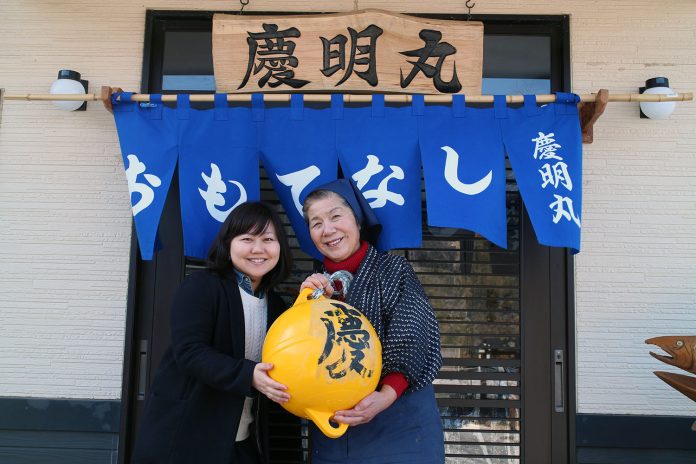 「慶明丸」と書かれた木の看板を掲げ、青い暖簾がかかった店の入り口前で、黄色い大きなウキを2人で抱えて笑顔で立っているリポーターの女性と三浦さんの写真