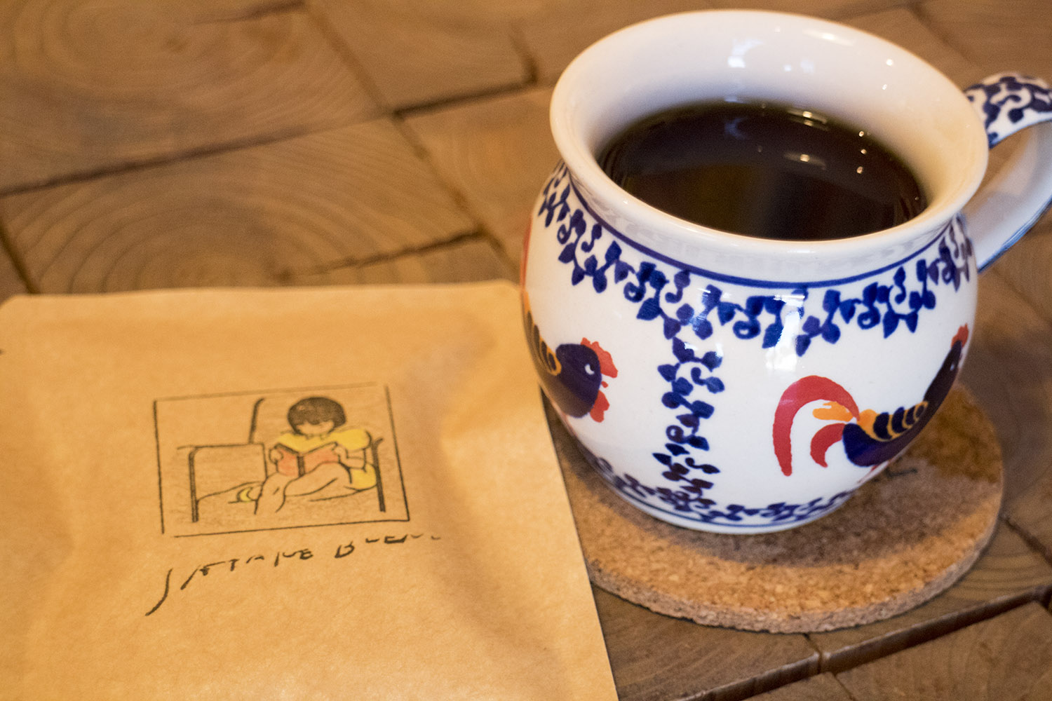 本を読む女の子のイラストが描かれた茶色のパッケージと鶏の絵が描かれた丸みを帯びたコーヒーカップが並べて置かれている写真