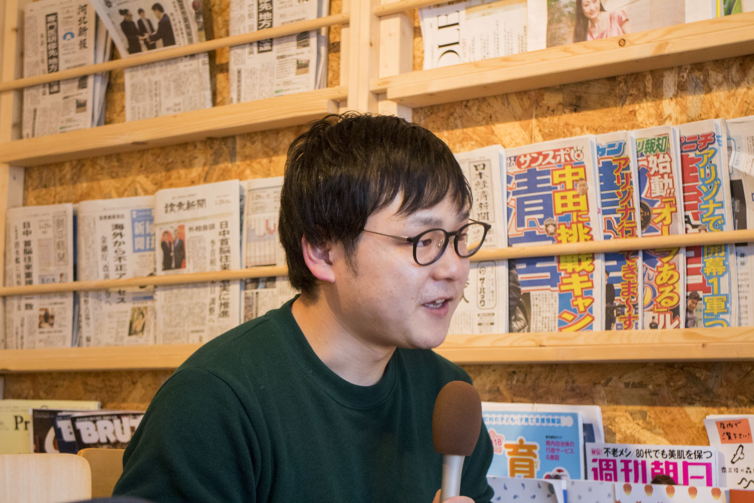 壁付のマガジンラックに様々な新聞、雑誌が並んだ前で、マイクを持ってインタビューを受けている長井さんの写真