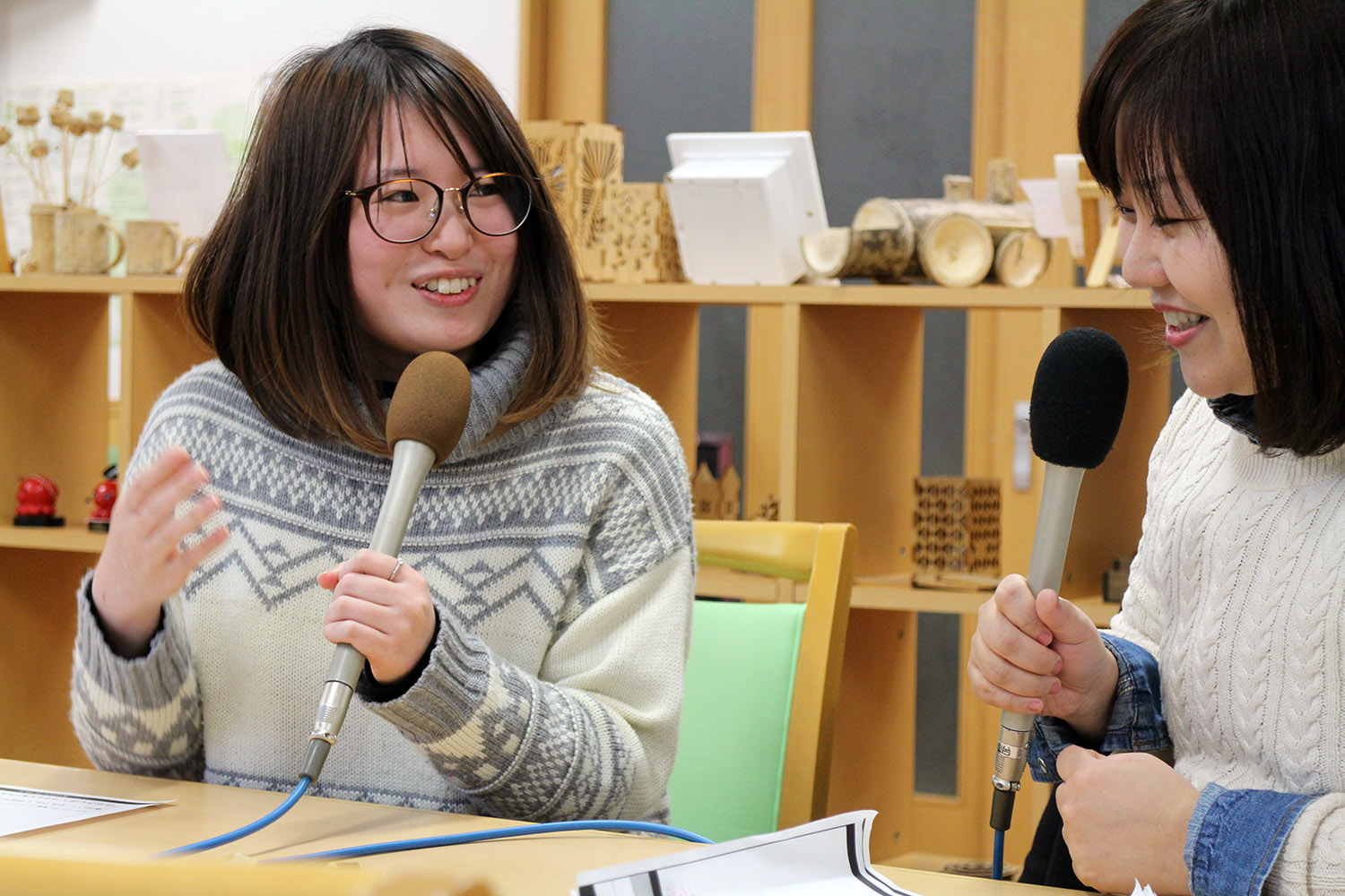 木の作品などが並ぶ木の棚の前で、佐藤さんとリポーターの女性がマイクを持ちながら楽しそうに会話している写真