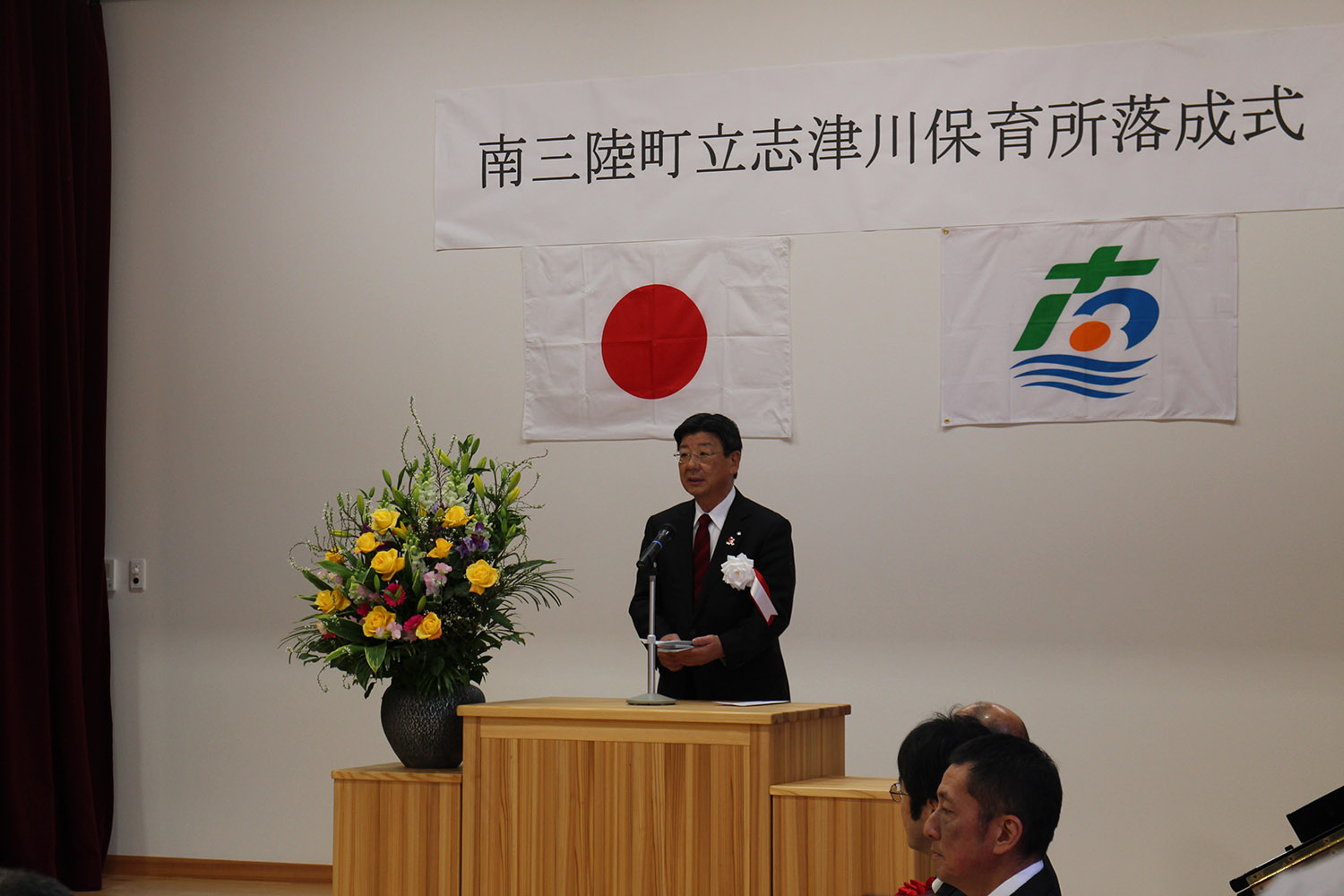 「南三陸町立志津川保育所落成式」と書かれた横断幕と日本国旗、町の旗を背に、壇上でスーツ姿の佐藤仁町長がマイクの前に立っている落成式の写真