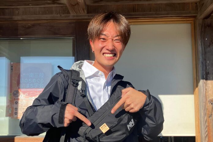 木造の建物の前に立っている星空之介さんが黒いショルダーバッグを指さしている写真