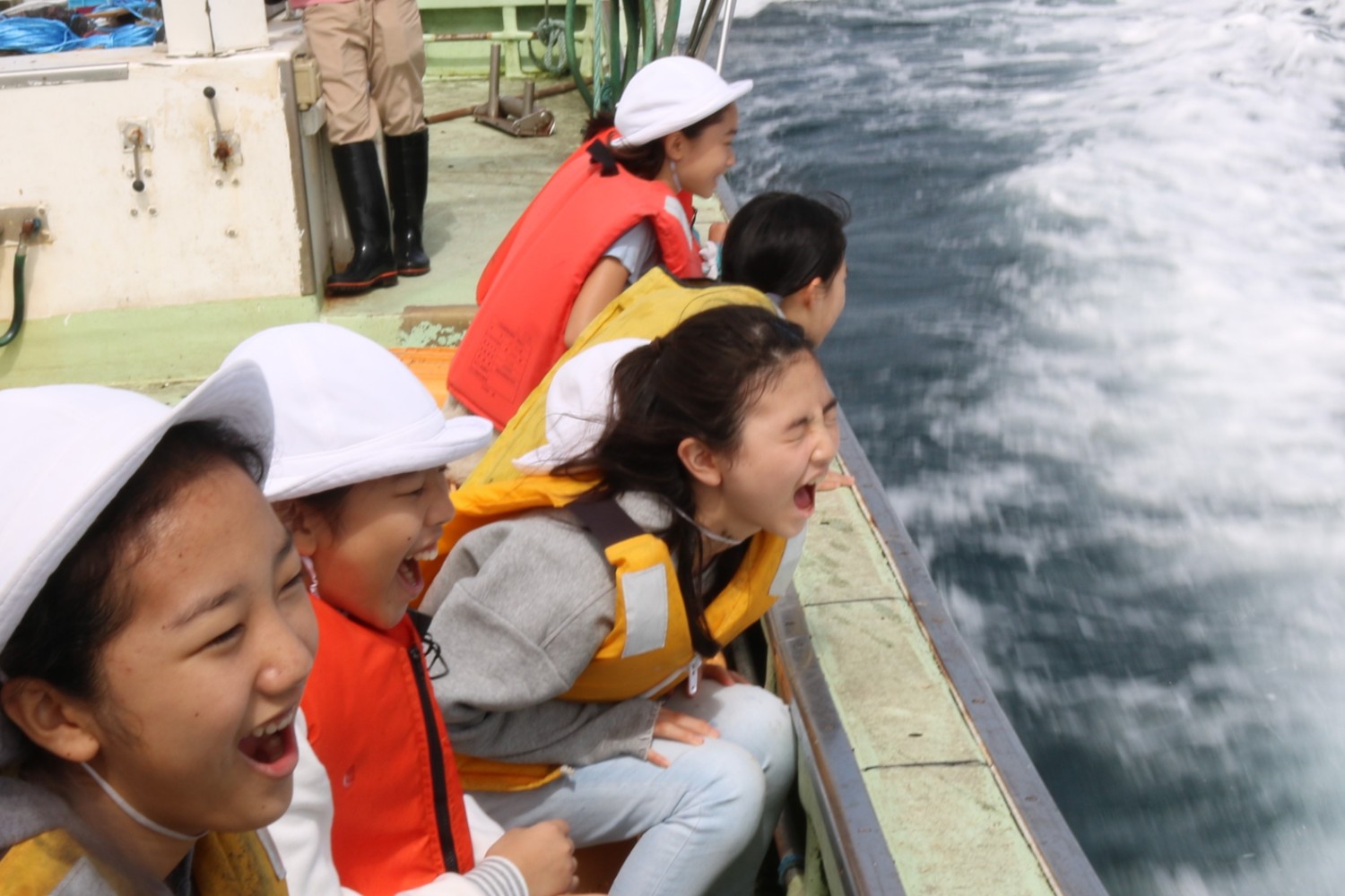救命胴衣を着た小学生たちが波を立てながら進む船の縁に身を乗り出して海面を見ながら歓声を上げて笑っている写真