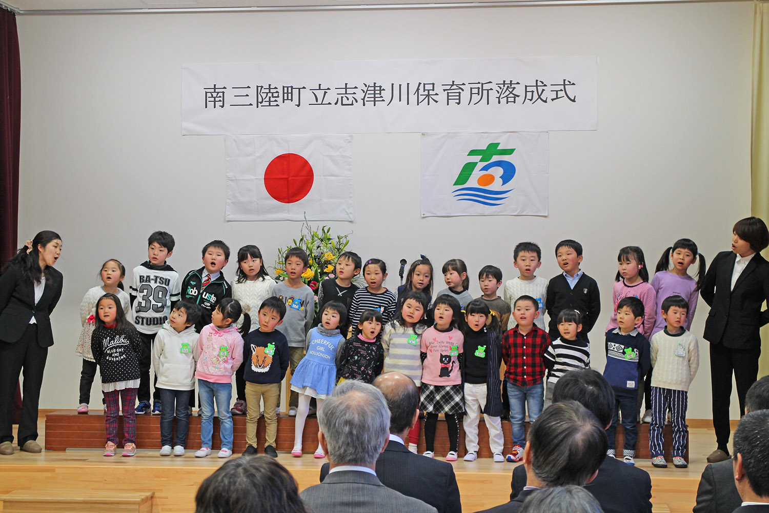 「南三陸町立志津川保育所落成式」と書かれた横断幕の前で、たくさんの園児たちがステージに並び、先生に見守られながら元気に歌をうたっている落成式の写真