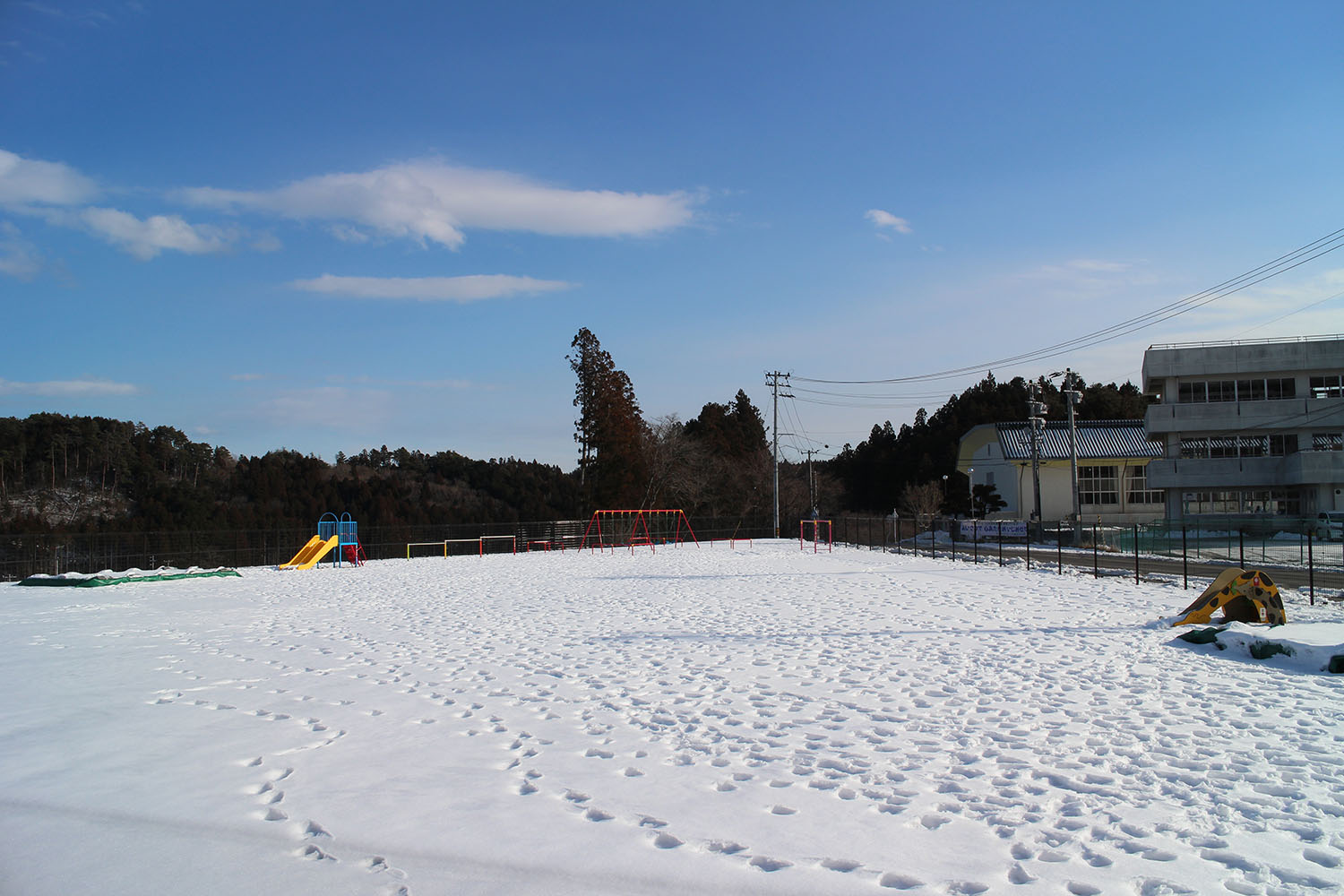雪に覆われた広い園庭に、滑り台やブランコなどの遊具が並び、奥に山と校舎が見える、志津川保育所の冬の園庭の風景を写した写真