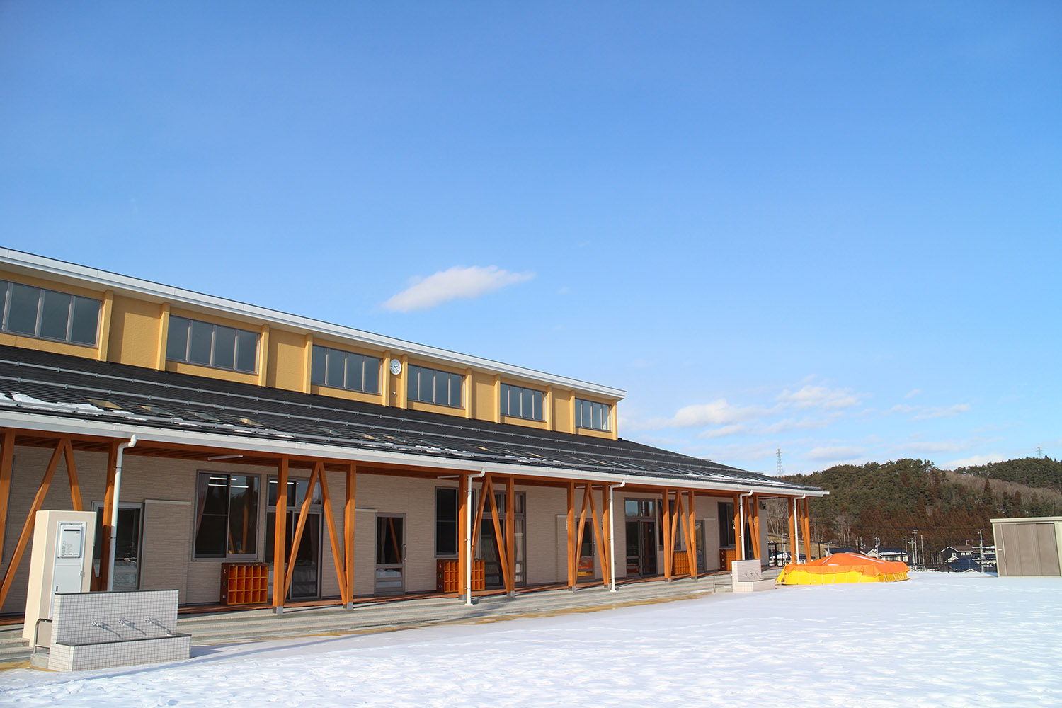 建物は黄色い上階と大きな窓が特徴的で、黒い屋根の下に木製の柱が等間隔に並ぶ横長の園舎で、広々とした園庭には真っ白い雪が積もっている志津川保育所の外観の写真