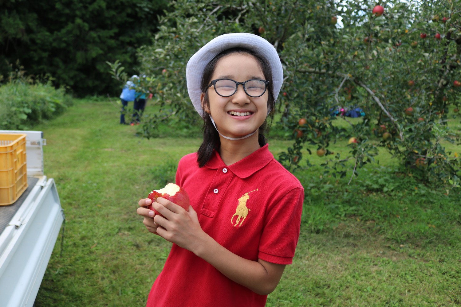 りんごの木と農園の緑が広がっている屋外で、白い帽子と赤いポロシャツを着た女の子がかじったリンゴを手に持って笑顔で立っている写真